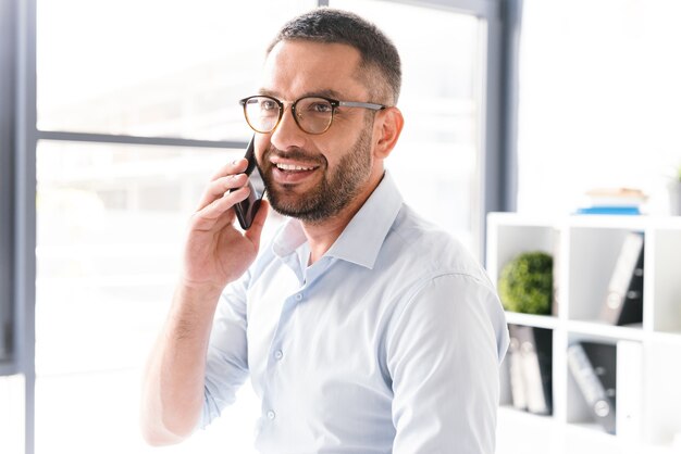 uomo allegro in abbigliamento formale e occhiali da vista parlando al telefono cellulare nero di affari, mentre si lavora in ufficio