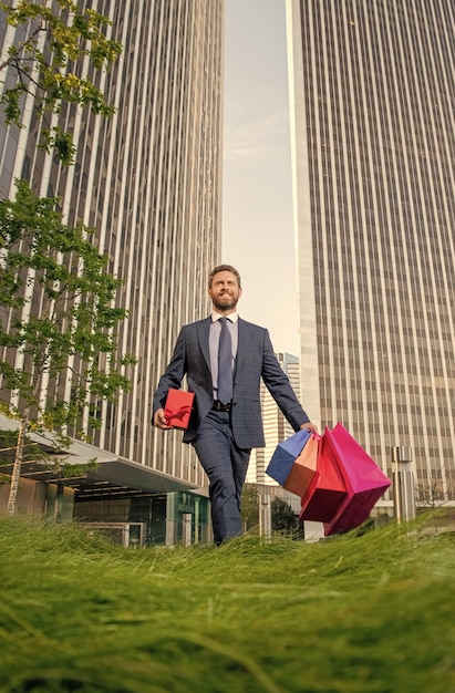Uomo allegro in abiti da cerimonia con borse della spesa e scatola regalo cammina fuori dal compleanno dell'ufficio