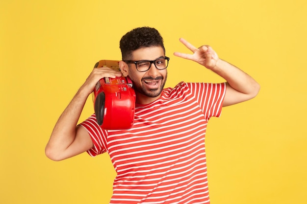 Uomo allegro sorridente con la barba in occhiali e maglietta a righe che tiene il giradischi sulla spalla e mostra il gesto di vittoria con le dita Colpo in studio al coperto isolato su sfondo giallo