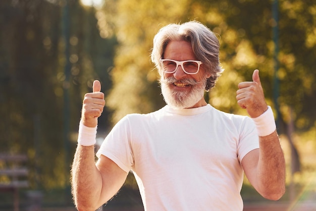 Uomo anziano alla moda in camicia bianca per occhiali e pantaloncini sportivi neri sul campo da tennis