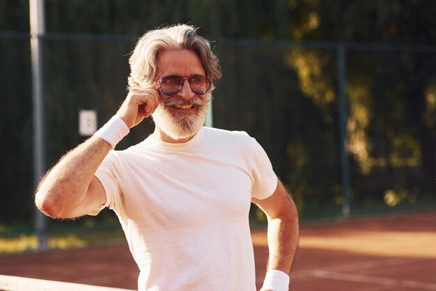 Uomo anziano alla moda in camicia bianca per occhiali e pantaloncini sportivi neri sul campo da tennis