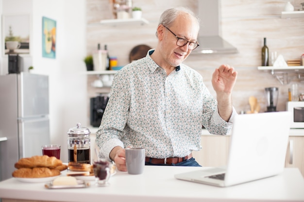 Uomo anziano allegro in videochiamata online dalla cucina mentre si gode la colazione e una tazza di caffè. Persona anziana che utilizza la webcam video con tecnologia di chat online su Internet che effettua una connessione videochiamata cam