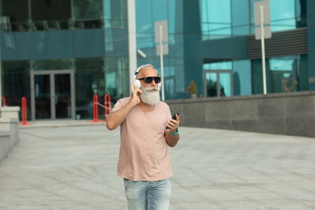 Uomo anziano anziano barbuto attraente e sorridente con capelli bianchi che gode della musica dalle cuffie bianche da portare all'aperto dello smart phone.