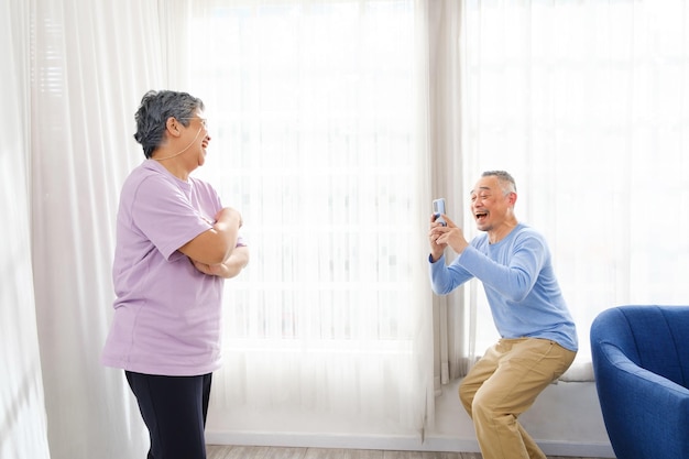 Uomo anziano asiatico in piedi con le ginocchia piegate a scattare foto di sua moglie con uno smartphone con un sorriso rilassato e ridere Attività indoor per anziani e pensionati