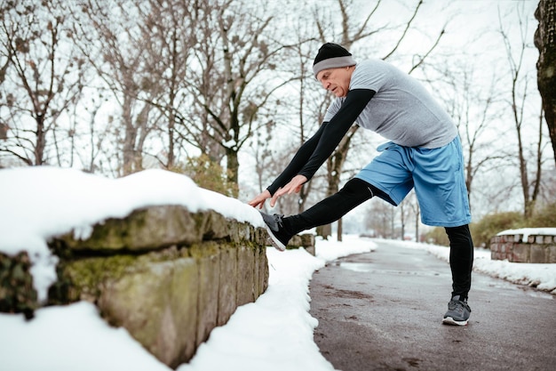 Uomo anziano attivo che si allunga e fa esercizi prima di fare jogging, nel parco pubblico durante l'allenamento invernale all'esterno. Copia spazio.