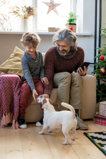 Uomo anziano barbuto e il suo simpatico nipotino che giocano con il cane mentre sono seduti in poltrona da abeti decorati contro la finestra alla vigilia di Natale