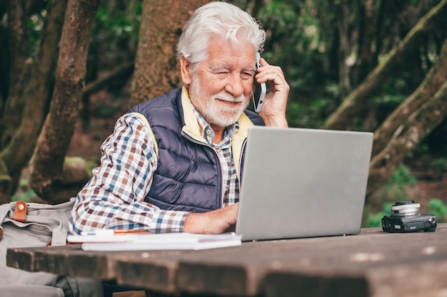 Uomo anziano barbuto sorridente che lavora al computer portatile utilizzando il telefono mentre si siede a un tavolo di legno in un parco