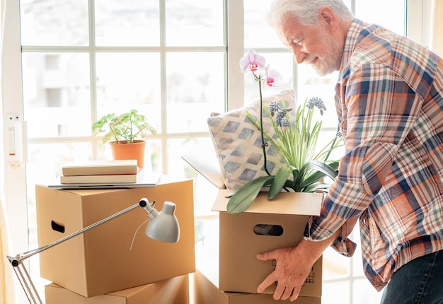 Uomo anziano bello sorridente che tiene scatola di cartone in posa nella nuova casa nel concetto di giorno commovente di trasferimento acquisto affitto casa appartamento