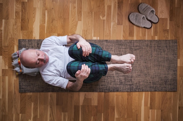 Uomo anziano caucasico in Apanasana o posa del vento a casa. Stile di vita sano e benessere, ricreazione, concetto di sport