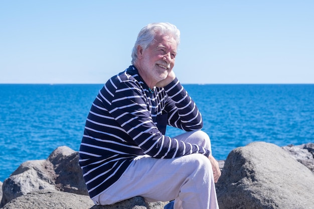 Uomo anziano caucasico sorridente seduto sulle scogliere che si gode il mare e la libertà in vacanza