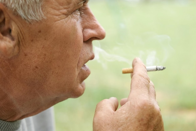 Uomo anziano che accende una sigaretta all'aperto sullo sfondo verde della natura Vecchio uomo premuroso che fuma guardando da parte Close Up Face Pensionato rilassato goditi il riposo del fine settimana fuori Abitudini malsane