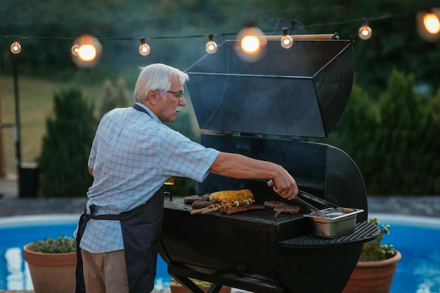 Uomo anziano che griglia carne al barbecue all'aperto con l'aiuto della pinza per barbecue