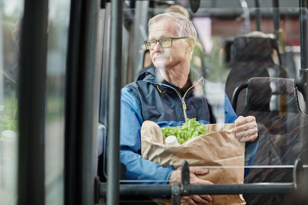 Uomo anziano che guarda il finestrino dell'autobus mentre viaggia con i mezzi pubblici in città e tiene in mano un ba...