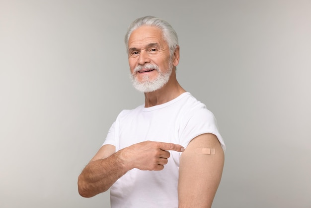 Uomo anziano che indica una benda adesiva dopo la vaccinazione su uno sfondo grigio chiaro