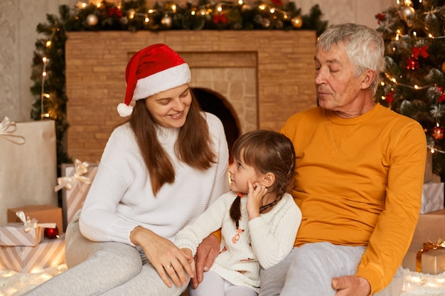 Uomo anziano che indossa un maglione arancione seduto con la sua famiglia vicino all'albero di Natale al coperto, persone che trascorrono il capodanno insieme, godendosi le vacanze invernali. Buon Natale e Felice Anno nuovo.