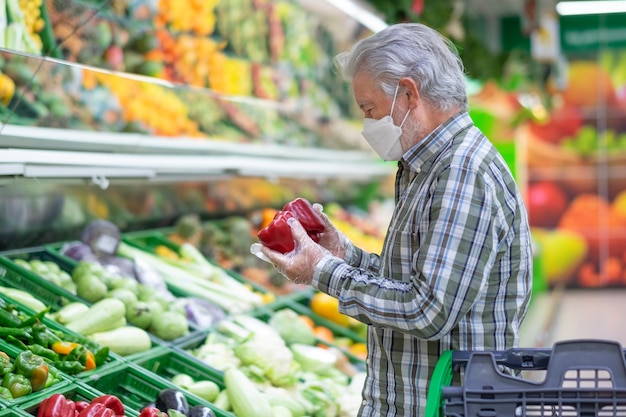Uomo anziano che indossa una maschera per evitare il contagio da covid 19 che fa shopping al supermercato selezionando le verdure