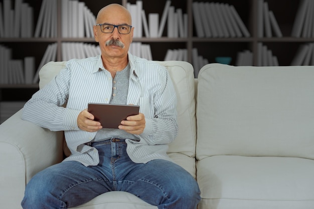 Uomo anziano che legge in una tavoletta digitale seduto sul divano del soggiorno di casa