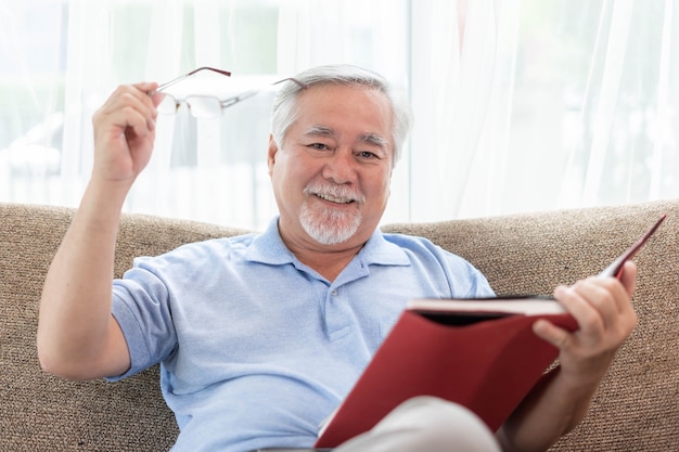 Uomo anziano che legge un romanzo libro, sorridente si sente felice sul divano di casa - concetto di anziani anziani di stile di vita