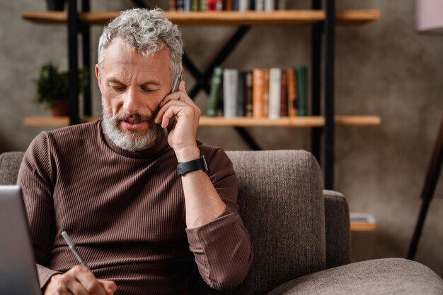 Uomo anziano che parla casualmente sul telefono cellulare e scrive note mentre lavora a casa utilizzando il laptop