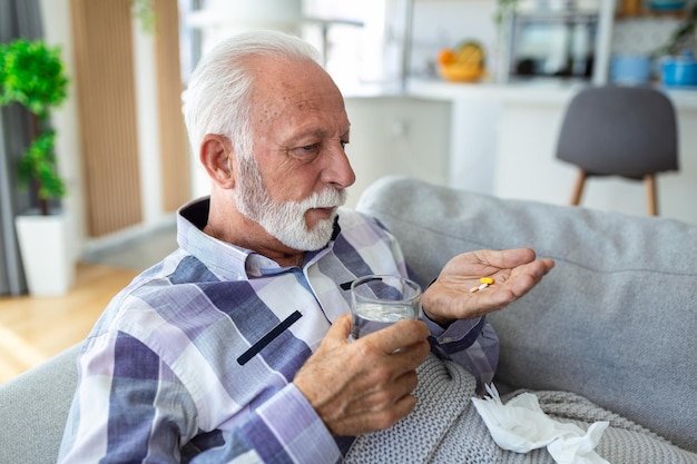 Uomo anziano che prende le pillole mentre è seduto sul divano a casa con in mano un vasetto bianco con spazio per la copia del trattamento Uomo anziano dai capelli grigi che usa integratori o vitamine Stile di vita sano nel concetto di età avanzata