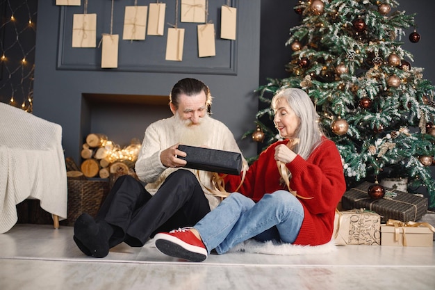 Uomo anziano che presenta il regalo di natale a sua moglie seduta vicino all'albero di natale