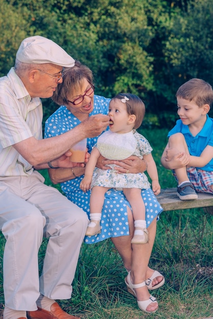 Uomo anziano che si alimenta con purea di frutta per una bambina adorabile seduta su una donna anziana in una panchina all'aperto. Concetto di stile di vita di nonni e nipoti.