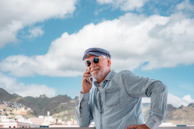 Uomo anziano che sorride e distoglie lo sguardo mentre usa il telefono vecchio che indossa occhiali da sole blu e berretto