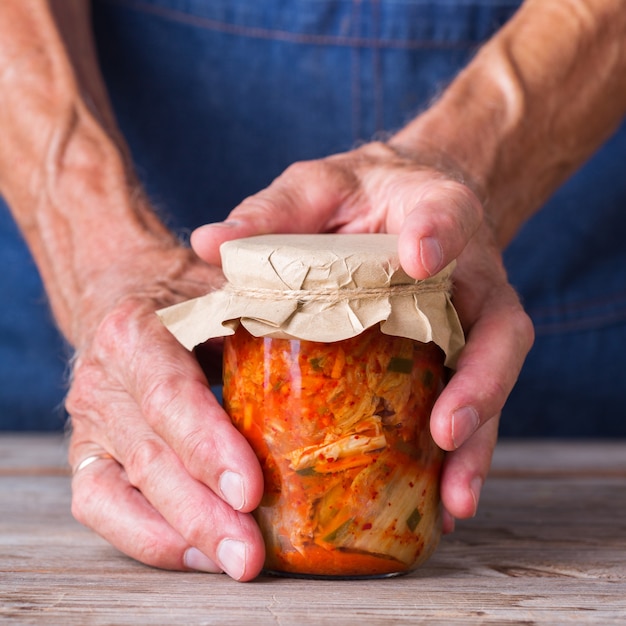 Uomo anziano che tiene in mano insalata di cavolo kimchi coreano tradizionale biologico fatto in casa in un barattolo di vetro su un tavolo di legno. Concetto di cibo salutare per l'intestino conservato vegetariano e vegano fermentato