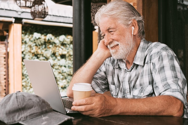 Uomo anziano che usa il computer guardando il video che indossa gli auricolari seduto all'aperto su un tavolo di legno Uomo anziano bello che naviga in internet sul computer portatile mentre beve un caffè