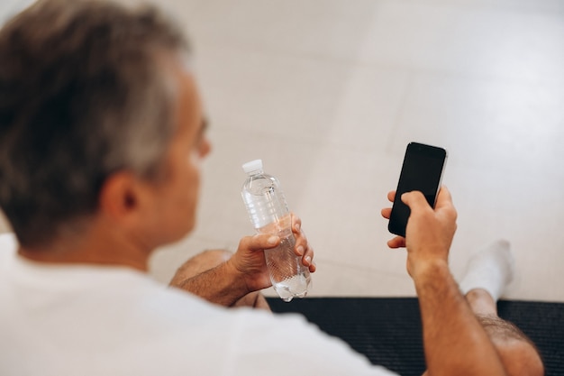 Uomo anziano che usa il suo smartphone e tiene in mano una bottiglia d'acqua di plastica