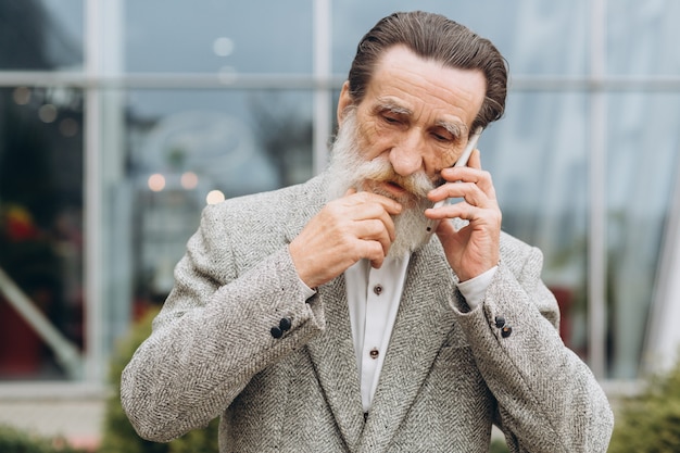 Uomo anziano con barba grigia e baffi in una giacca parla al telefono