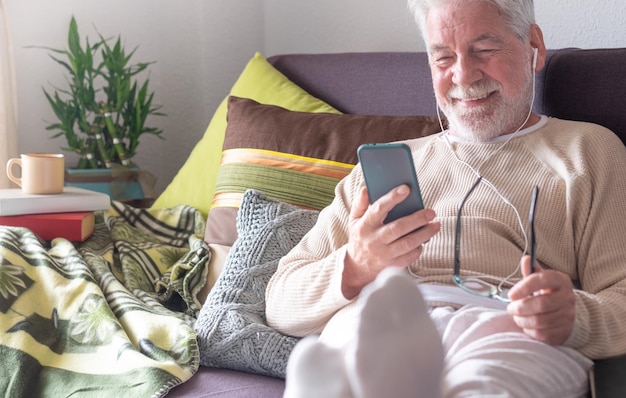 Uomo anziano dai capelli bianchi bello con gli auricolari sdraiato sul divano di casa utilizzando lo smartphone Luce intensa dalla finestra Anziani in pensione che utilizzano la tecnologia wireless
