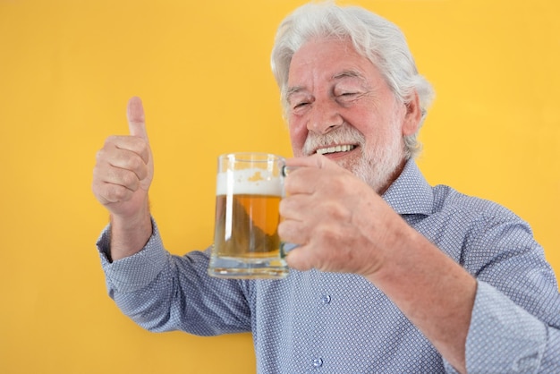 Uomo anziano dai capelli bianco felice che tiene un bicchiere di birra alcolica fresca con il pollice in su