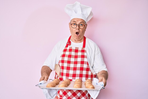 Uomo anziano dai capelli grigi che indossa l'uniforme da panettiere che tiene il pane fatto in casa impaurito e scioccato dalla sorpresa e dall'espressione stupita paura e faccia eccitata