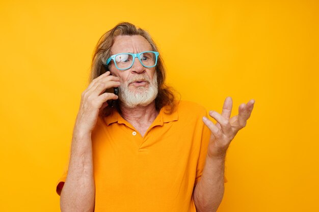 Uomo anziano dai capelli grigi che indossa una camicia gialla con occhiali blu che parla al telefono sullo sfondo isolato