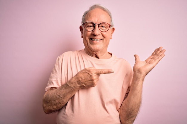 Uomo anziano dai capelli grigi con gli occhiali in piedi su sfondo rosa isolato stupito e sorridente alla telecamera mentre si presenta con la mano e indica con il dito