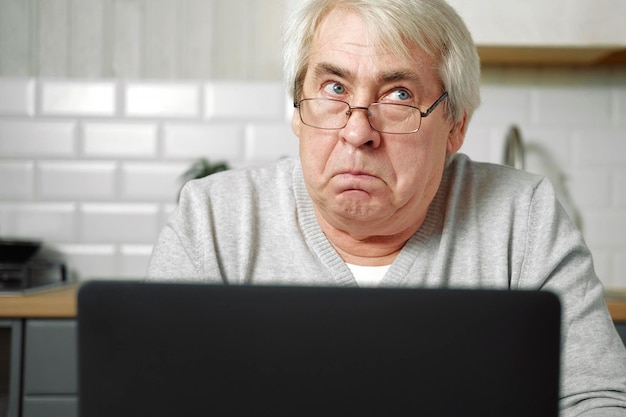 Uomo anziano dai capelli grigi con gli occhiali seduto al computer portatile e che fa la faccia di pesce con le labbra Gesto pazzo e comico Divertente vecchio nonno con un'espressione facciale sciocca e giocosa che fa smorfie scherzando