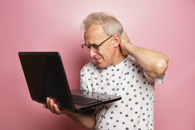 Uomo anziano dai capelli grigi con una maglietta bianca che tiene un laptop nero e si gratta la parte posteriore della testa su uno sfondo rosa, generazione senior e concetto di tecnologia moderna