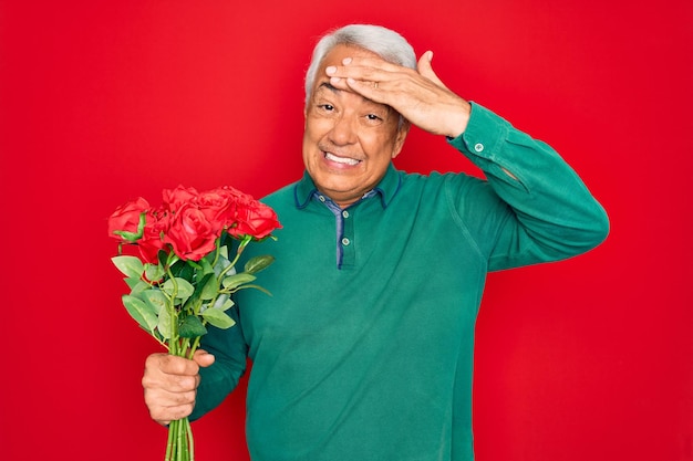 Uomo anziano dai capelli grigi di mezza età che tiene un romantico bouquet di rose rosse sullo sfondo con la mano sulla testa