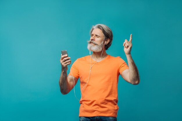 Uomo anziano dai capelli grigi in cuffie cablate con un telefono in posa su sfondo blu Riprese in studio