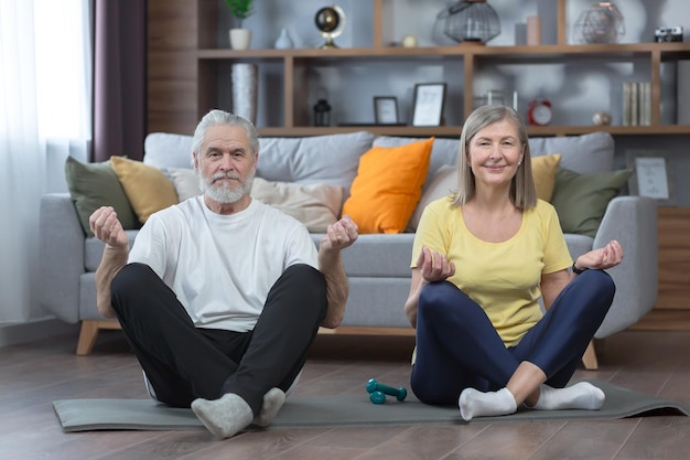 Uomo anziano e donna in casa che fanno yoga in famiglia su una stuoia nella posizione del loto guardando la telecamera insieme esercizio mattutino e fitness