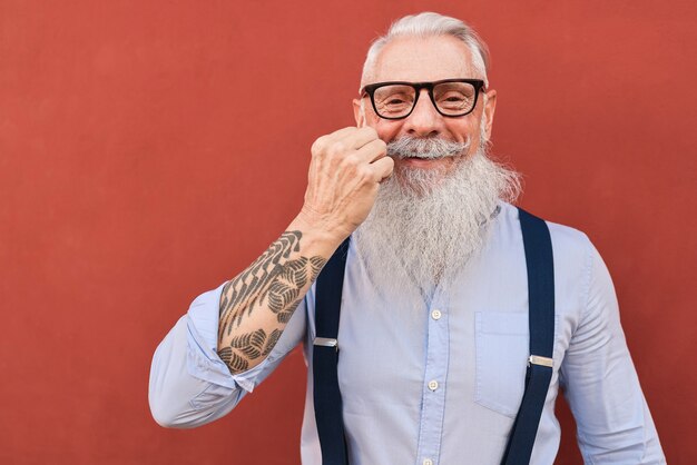 Uomo anziano felice hipster che tocca i baffi con la parete rossa