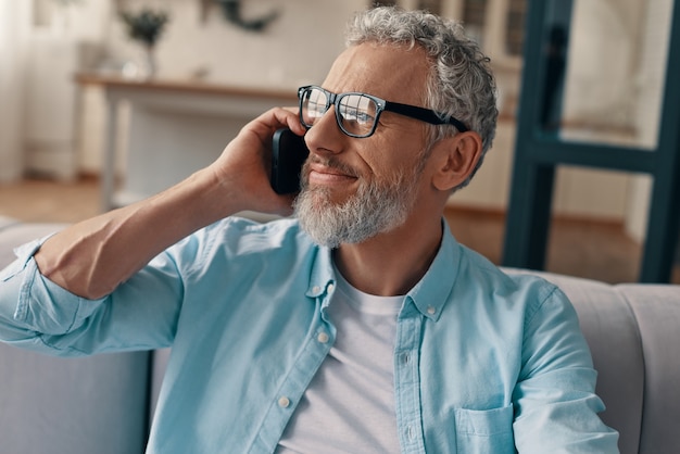 Uomo anziano in abbigliamento casual e occhiali che parla allo smartphone mentre è seduto sul divano di casa