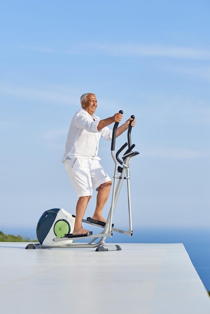 uomo anziano in buona salute che si allena sulla macchina del tapis roulant della palestra nella moderna terrazza domestica con vista sull'oceano