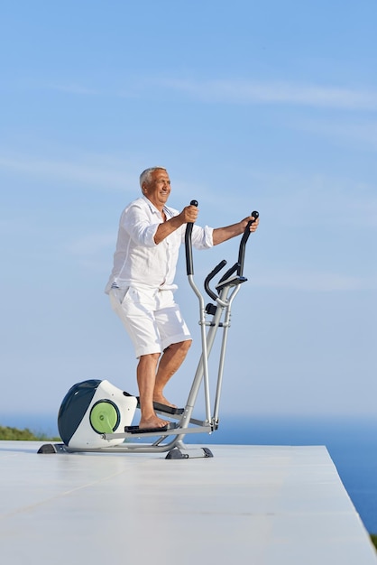 uomo anziano in buona salute che si allena sulla macchina del tapis roulant della palestra nella moderna terrazza domestica con vista sull'oceano