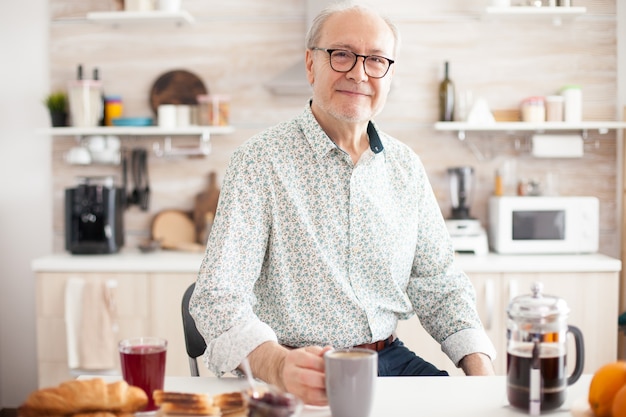 Uomo anziano in cucina che sorride guardando la telecamera tenendo una tazza di caffè caldo. Ritratto di una persona anziana anziana rilassata al mattino, gustando una bevanda calda fresca. Viso adulto sano e sorridente