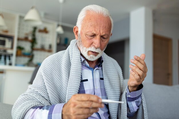 Uomo anziano malato che controlla la sua temperatura che soffre di influenza stagionale o raffreddore che giace sul divano che tossisce che soffre dell'influenza stagionale o del freddo che soffre l'anziano che si sente malato con l'influenza a casa