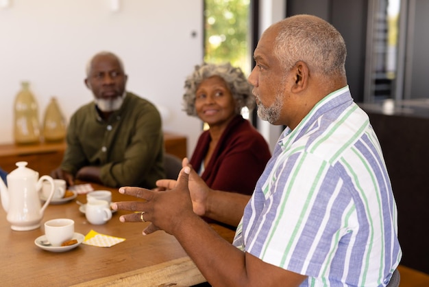 Uomo anziano multirazziale che parla con gli amici mentre beve caffè e biscotti al tavolo da pranzo. Casa di cura, bevande, cibo, inalterato, amicizia, solidarietà, sostegno, vita assistita e pensione.
