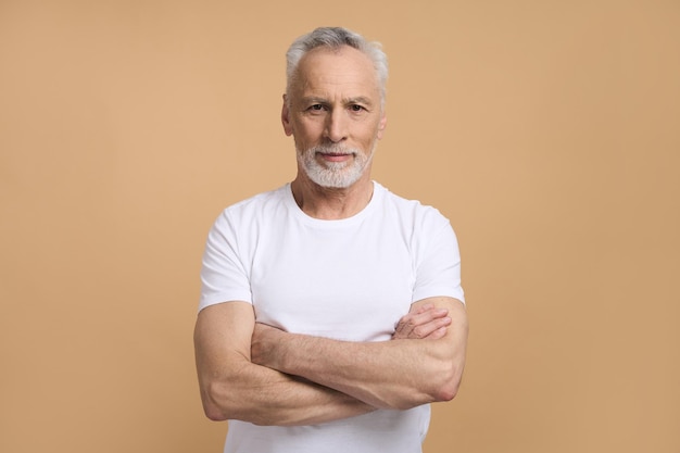 Uomo anziano pensionato caucasico che sorride alla macchina fotografica che posa con le armi piegate isolato su fondo beige