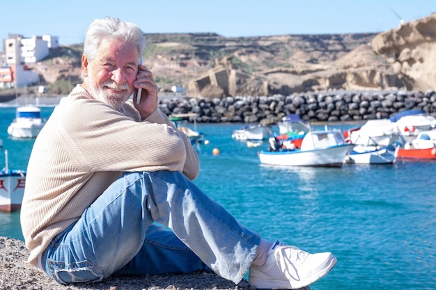Uomo anziano rilassato seduto al porto usando il suo smartphone. Attraenti anziani sorridenti che si godono la vita all'aria aperta e la bellezza della natura. Barche da pesca e costa sullo sfondo